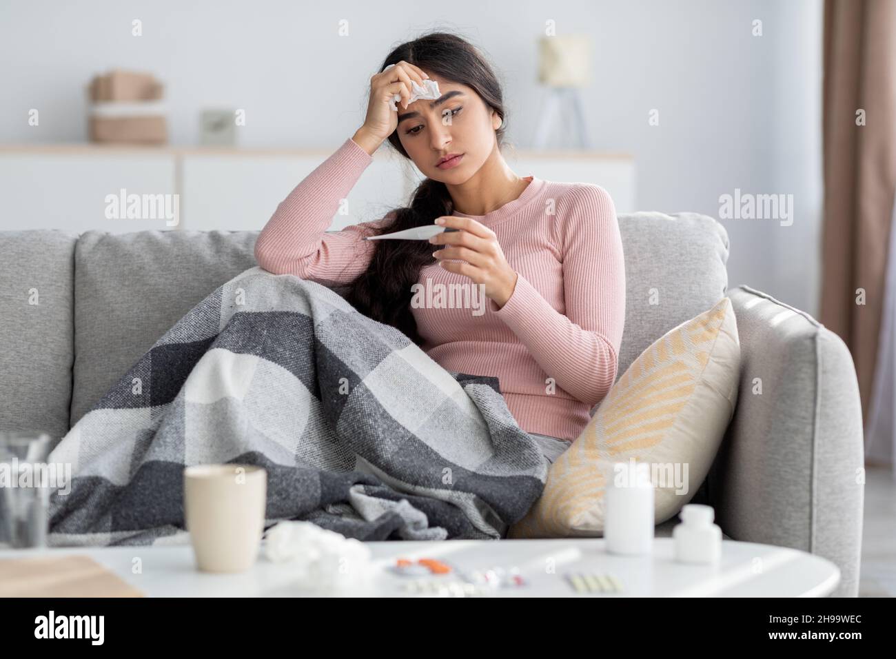 Jeune femme hindoue triste et découplée avec du plaid, souffre de la grippe, s'assoit sur un canapé et regarde le thermomètre dans le salon Banque D'Images