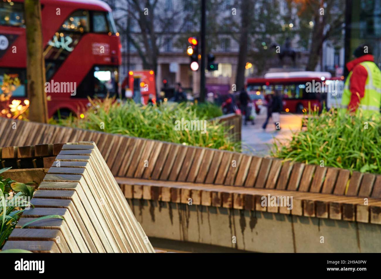 Scène de rue au crépuscule de Londres avec un arrière-plan flou avec bus de londres, piétons, plantes et sièges en bois. Aucune marque visible. Banque D'Images