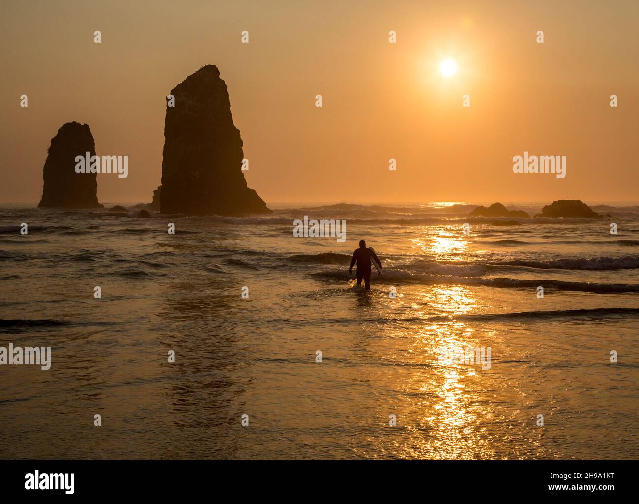 Surfeur marchant hors de la mer avec le soleil se coucher derrière le rocher dans les vagues sur la plage. Banque D'Images