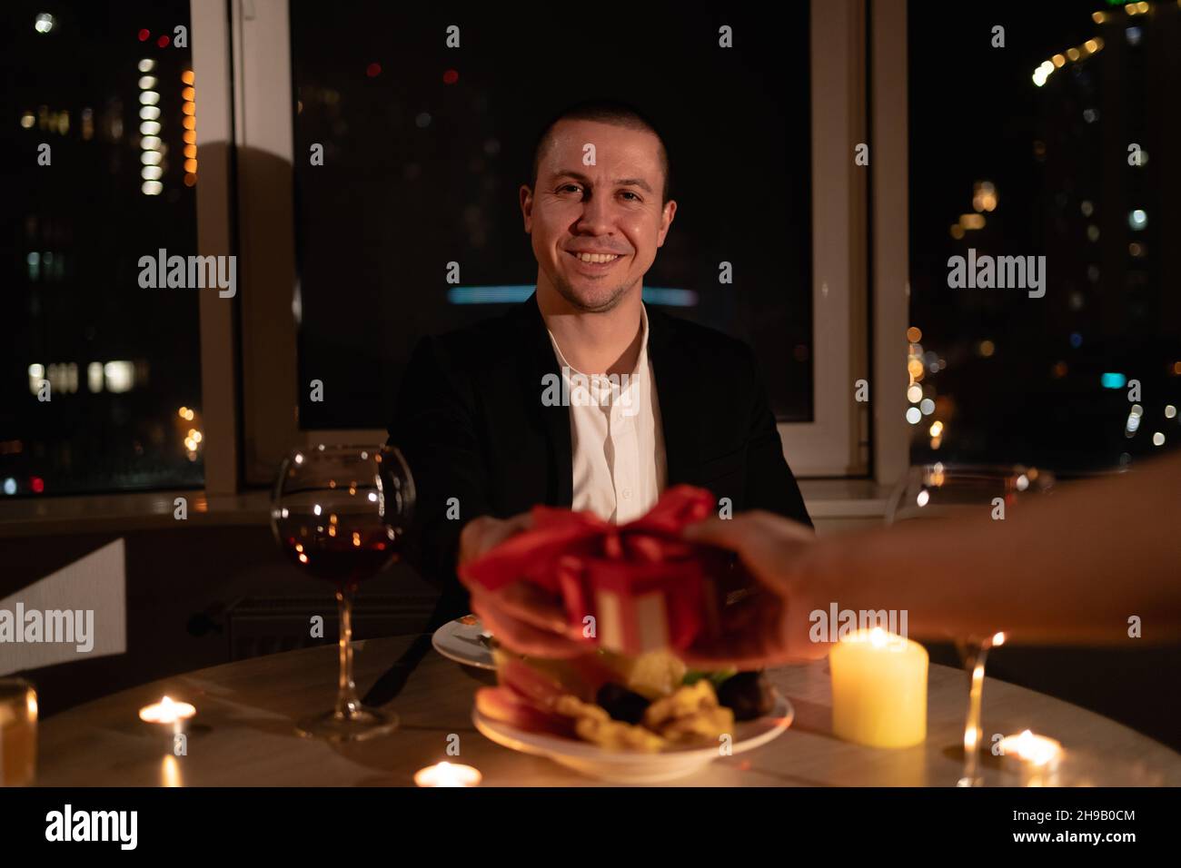 le jour de la saint-valentin aux chandelles, la main féminine donne un cadeau à un homme assis à la table, date romantique, portrait d'un homme amoureux en costume avec vin Banque D'Images