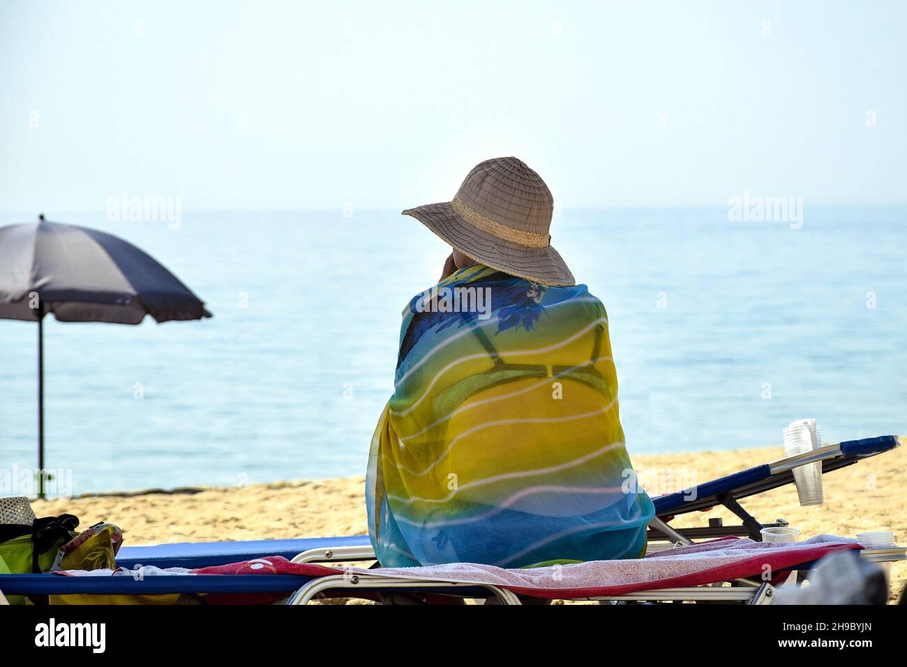 Vacances d'été.Détente sur la plage.Femme âgée en maillot de bain, pareo et panama assis sur une chaise longue sous un parapluie sur la plage et regarde int Banque D'Images