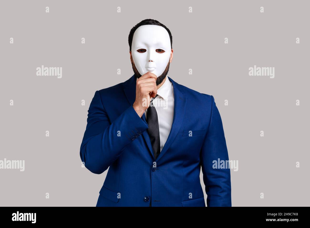 Homme d'affaires inconnu anonyme couvrant son visage avec un masque blanc, cachant sa vraie personnalité, l'anonymat, portant un costume de style officiel.Prise de vue en studio isolée sur fond gris. Banque D'Images