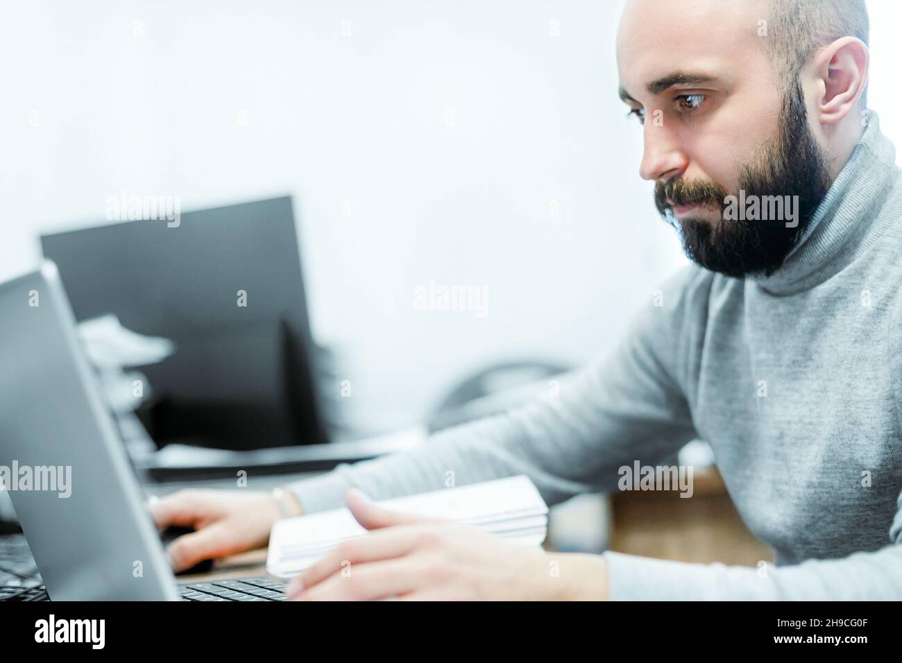 Un jeune homme à tête blanche et barbu de 25-35 ans travaille au bureau pour un ordinateur portable.Guy dans les vêtements décontractés regarde l'écran d'ordinateur portable.Scène réelle.Un vrai travailleur. Banque D'Images