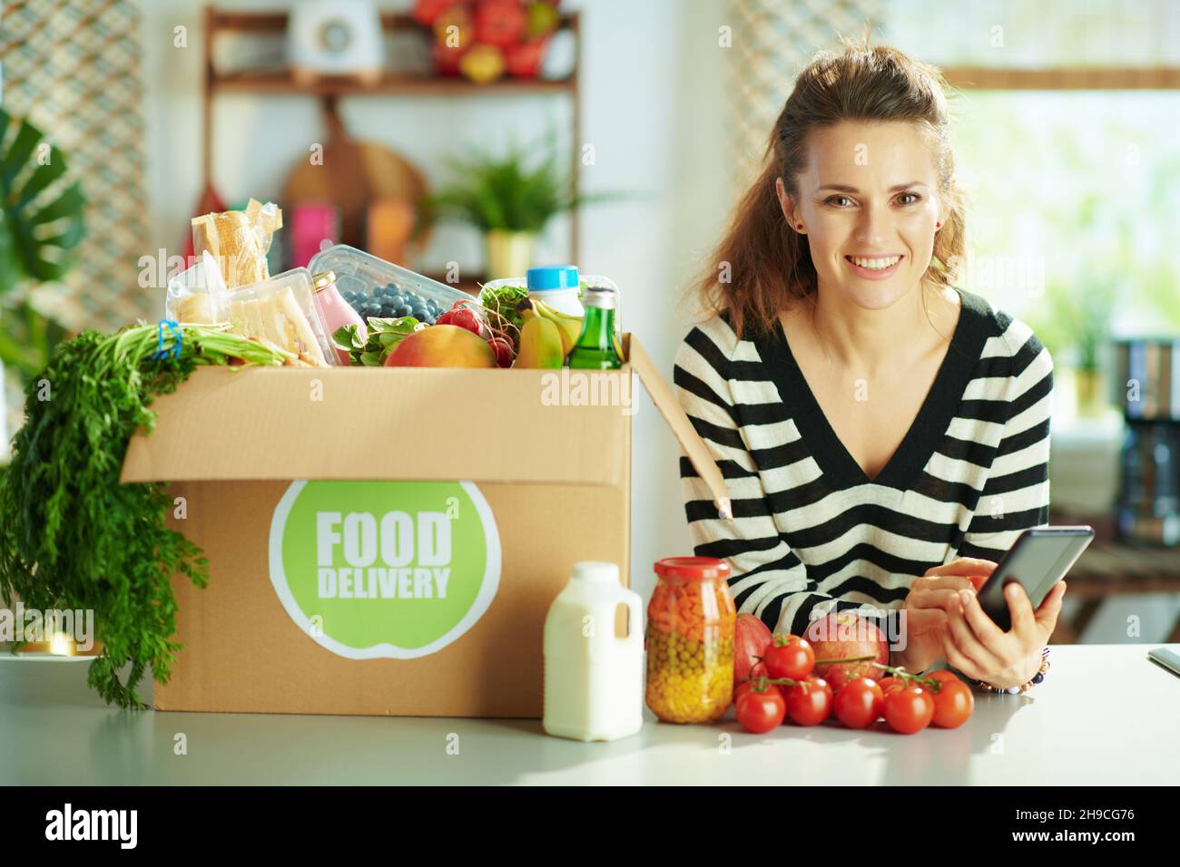 Livraison de nourriture. Femme de ménage souriante d'âge moyen avec boîte à nourriture à l'aide de l'application recettes de smartphone dans la cuisine. Banque D'Images