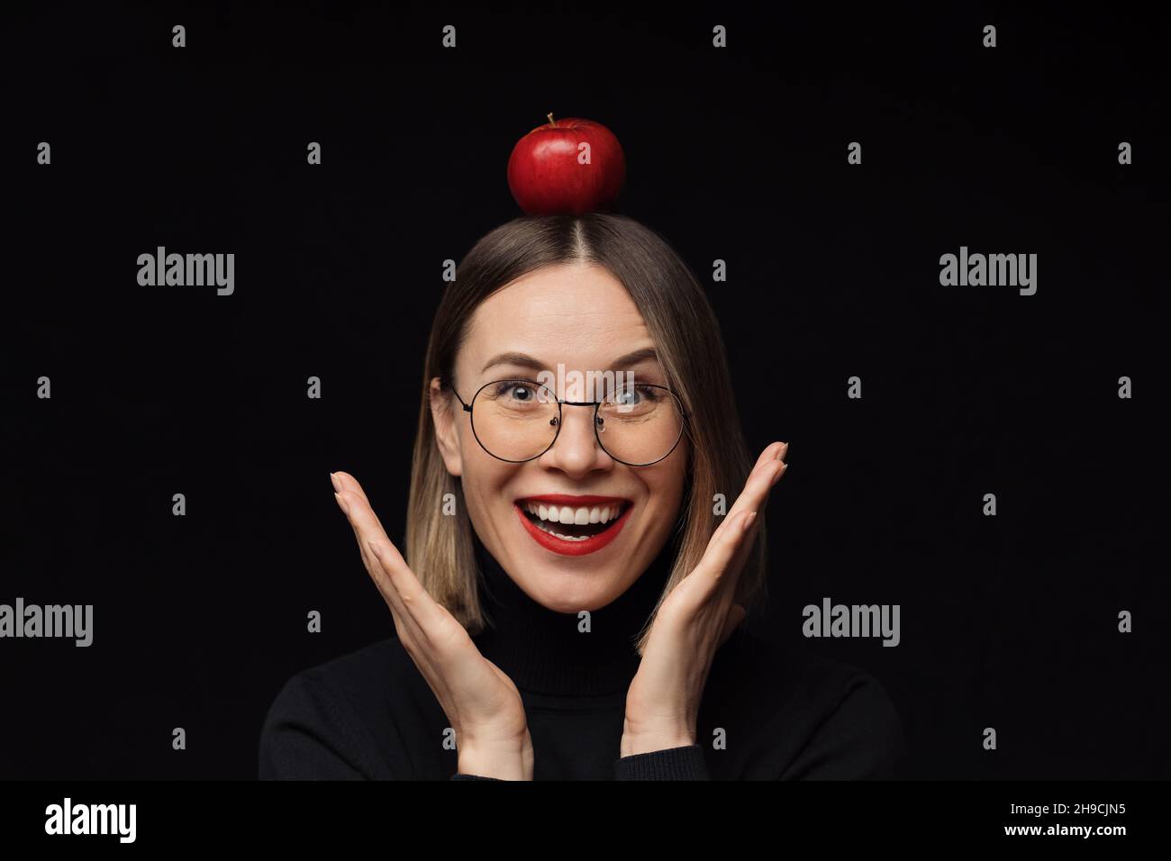 Gros plan surprise femme portrait portant des lunettes avec des cadres noirs, avec des lèvres rouges, regardant l'appareil photo avec une pomme rouge sur la tête sur fond noir. Banque D'Images