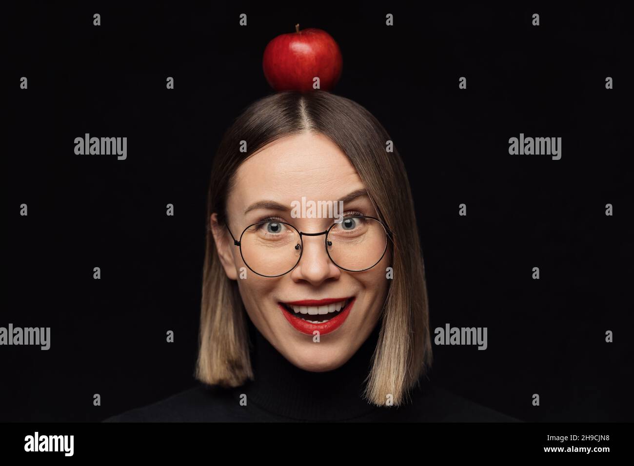 Gros plan surprise femme portrait portant des lunettes avec des cadres noirs, avec des lèvres rouges, regardant l'appareil photo avec une pomme rouge sur la tête sur fond noir. Banque D'Images