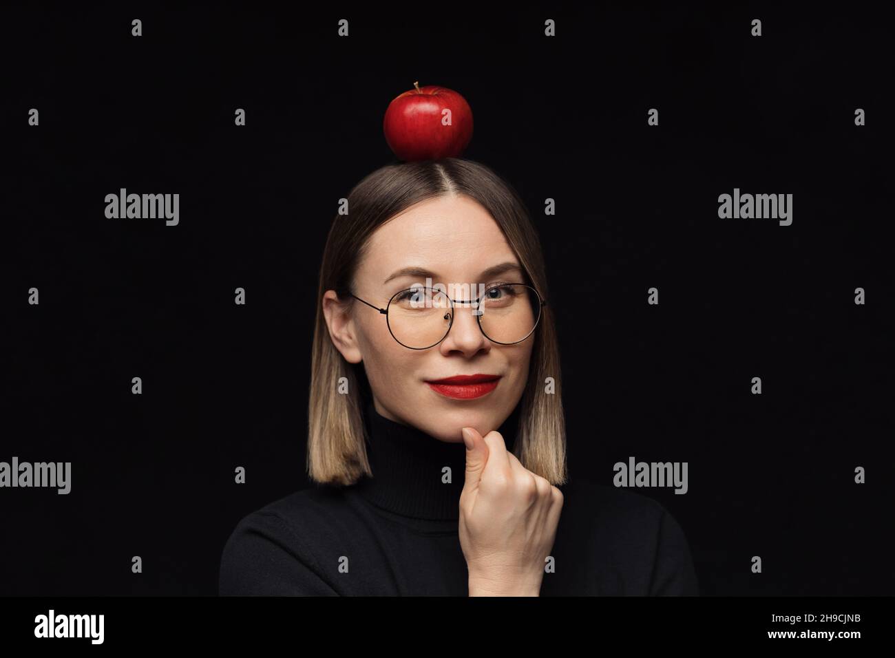 Gros plan sourire femme portrait portant des lunettes avec des cadres noirs, avec des lèvres rouges, regardant l'appareil photo avec une pomme rouge sur la tête sur fond noir maquette. Banque D'Images