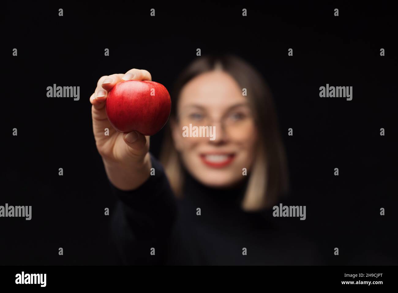 Une femme souriante portant des lunettes montre une pomme rouge à l'appareil photo, avec des lèvres rouges, sur un fond noir.Concentrez-vous sur les fruits Banque D'Images