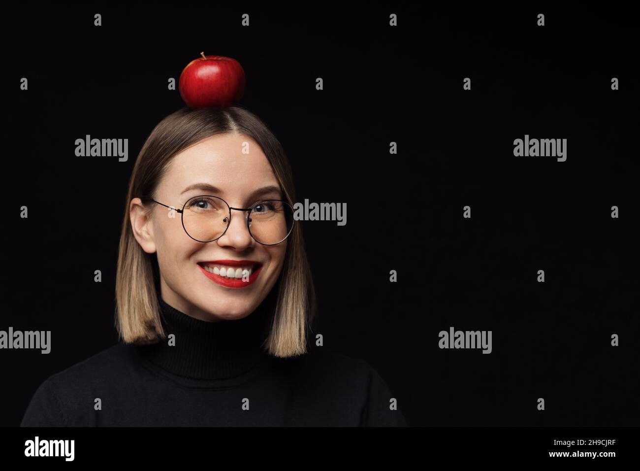 Gros plan sourire femme portrait portant des lunettes avec des cadres noirs, avec des lèvres rouges, regardant l'appareil photo avec une pomme rouge sur la tête sur fond noir maquette. Banque D'Images