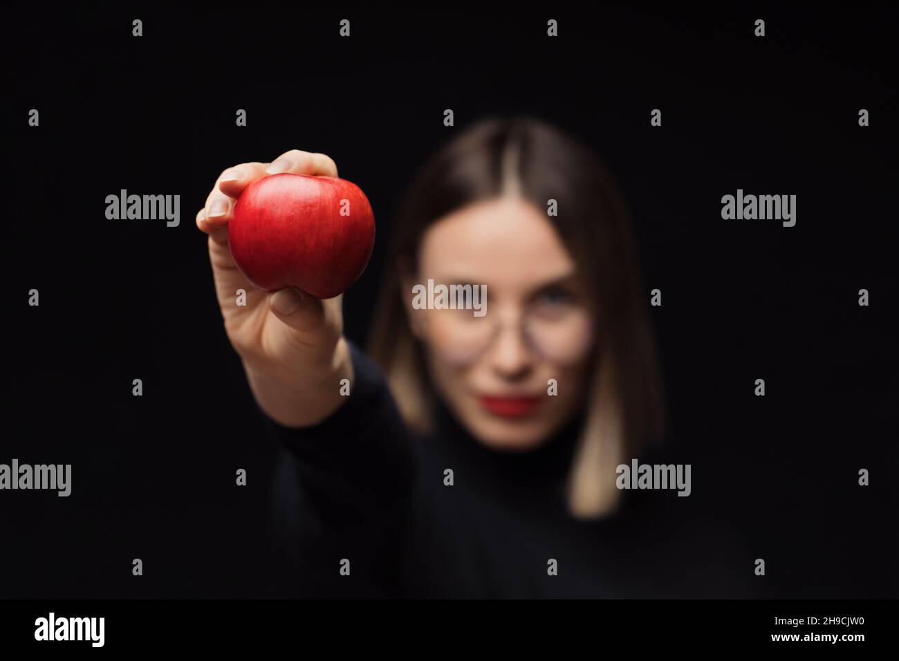 Une femme souriante portant des lunettes montre une pomme rouge à l'appareil photo, avec des lèvres rouges, sur un fond noir.Concentrez-vous sur les fruits Banque D'Images