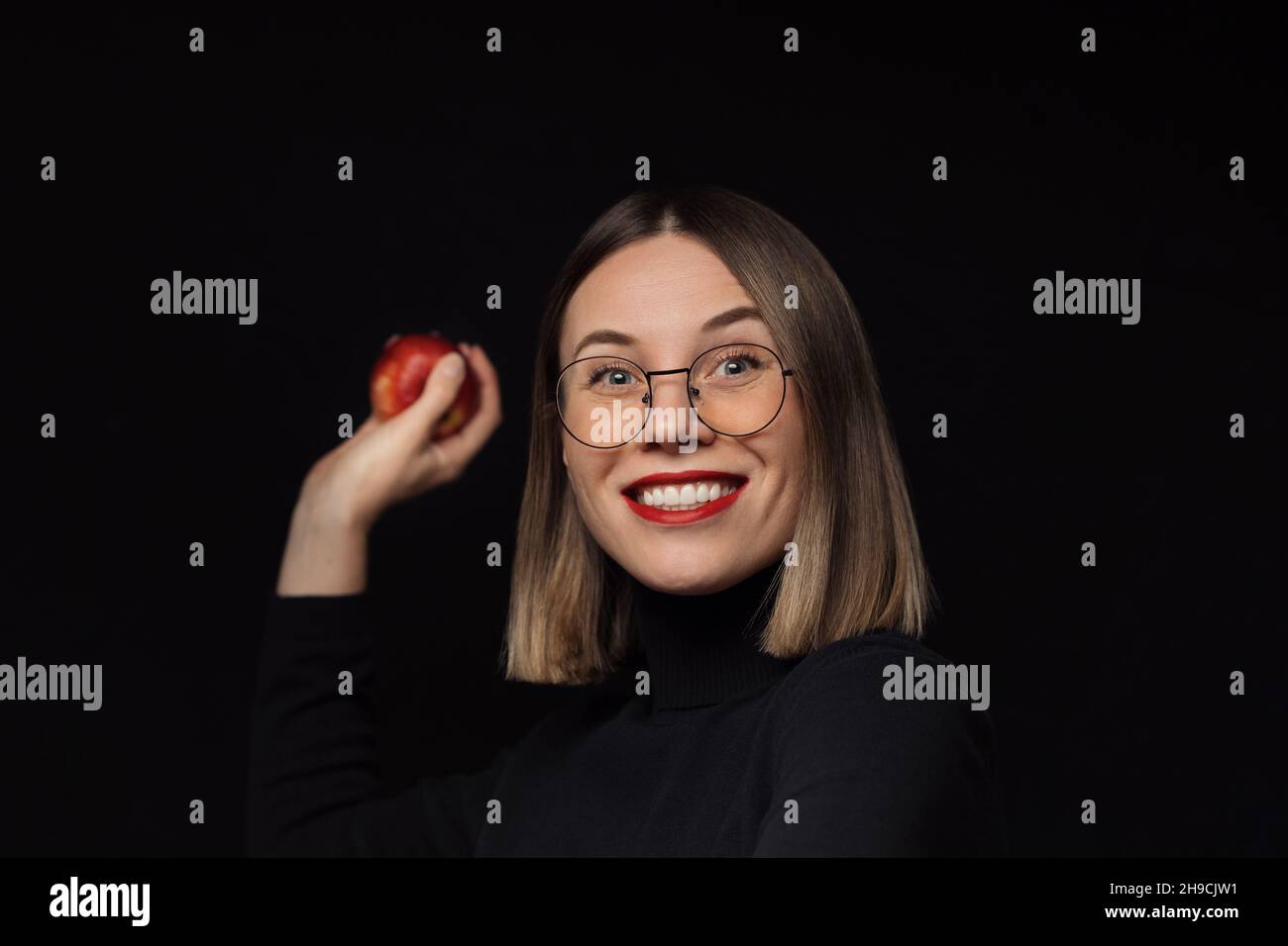 Femme excitée portant des lunettes avec des cadres noirs, avec des lèvres rouges, regardant l'appareil photo veut jeter une pomme rouge sur fond noir. Banque D'Images
