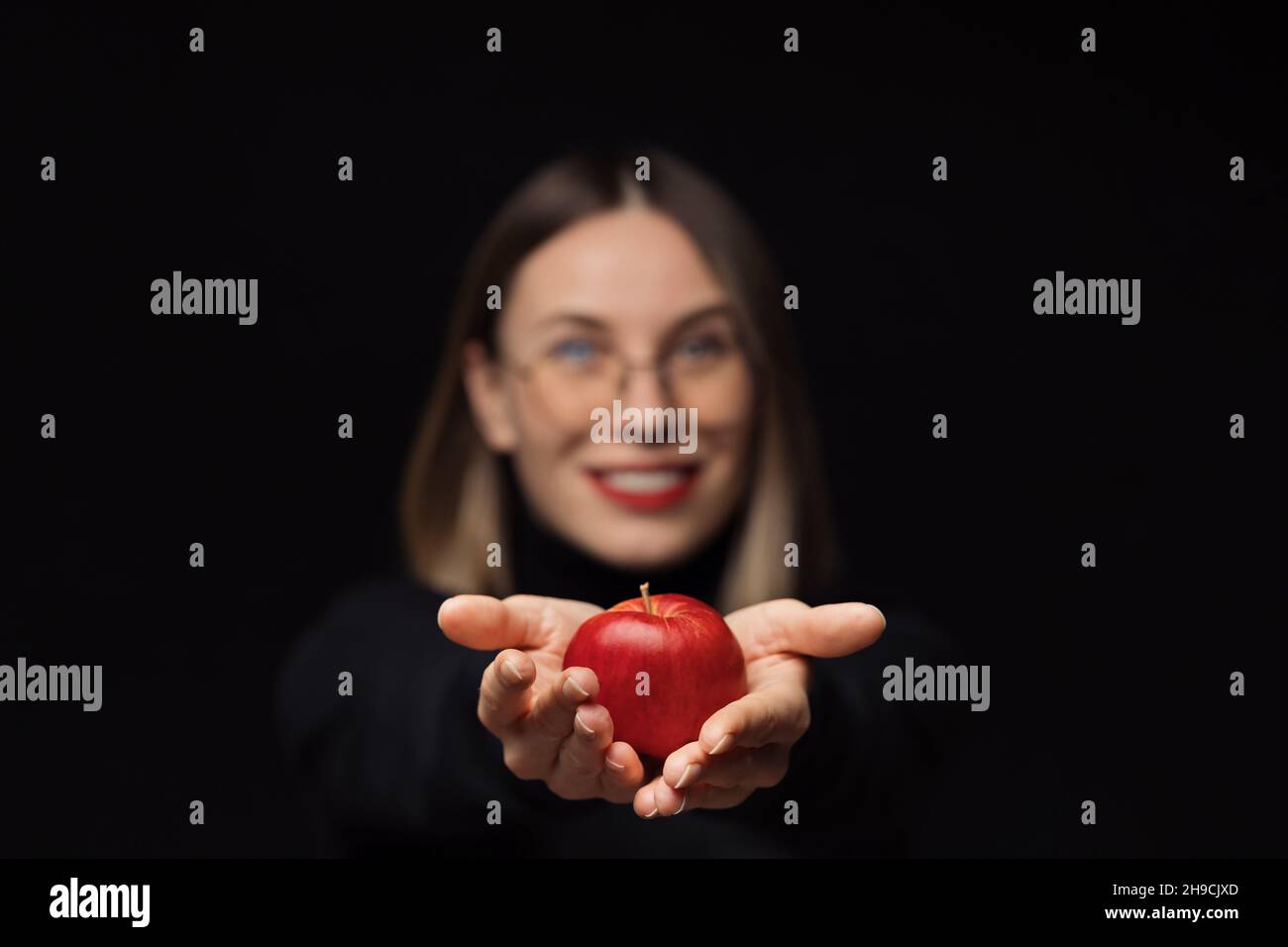 Une femme souriante portant des lunettes tenant une pomme rouge dans les palmiers la montre à l'appareil photo, avec des lèvres rouges, sur un fond noir.Concentrez-vous sur les fruits Banque D'Images