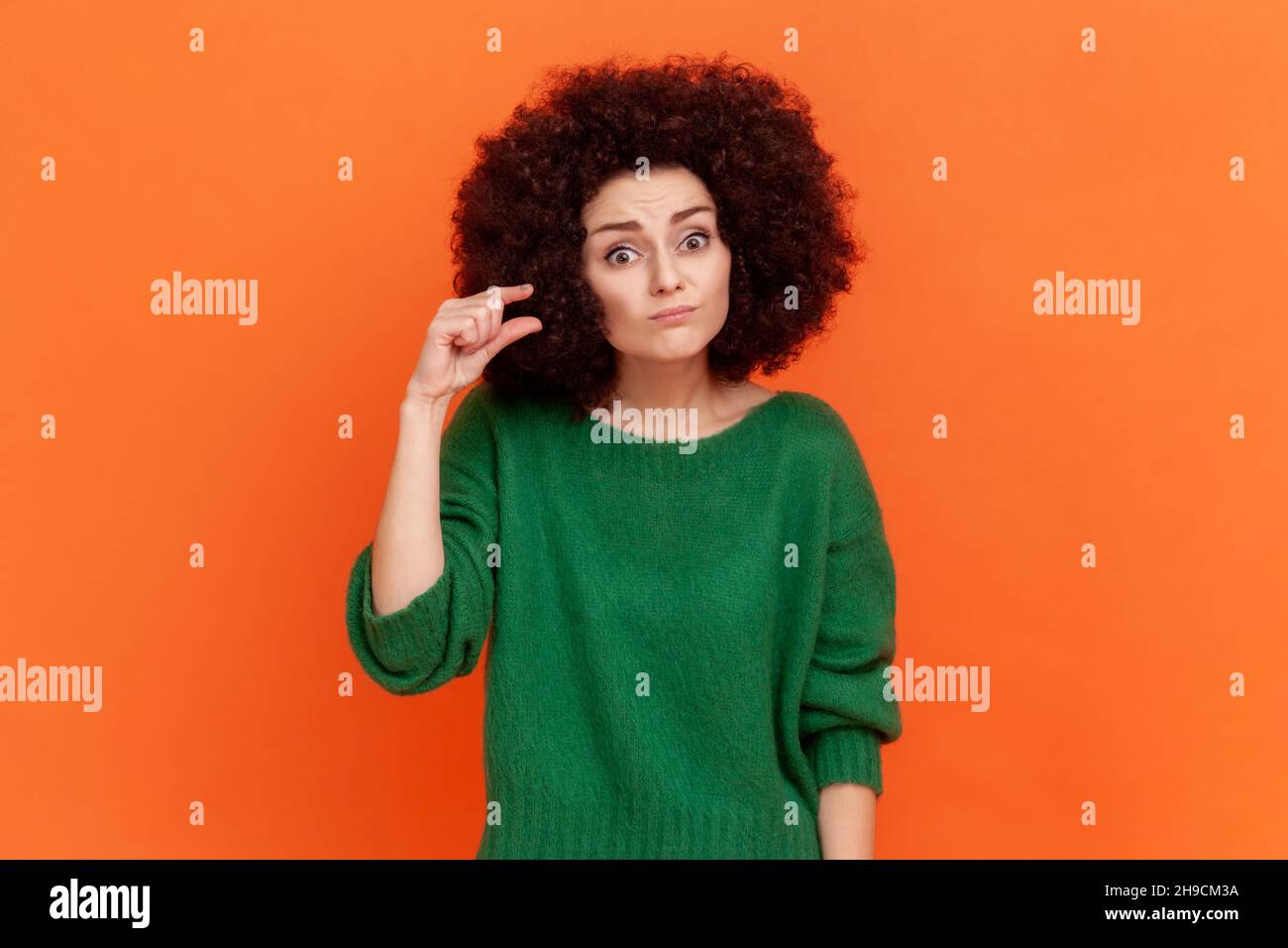 Portrait d'une femme avec une coiffure afro portant un chandail vert de style décontracté montrant un petit geste avec les doigts, regardant l'appareil photo avec déception.Studio d'intérieur isolé sur fond orange Banque D'Images