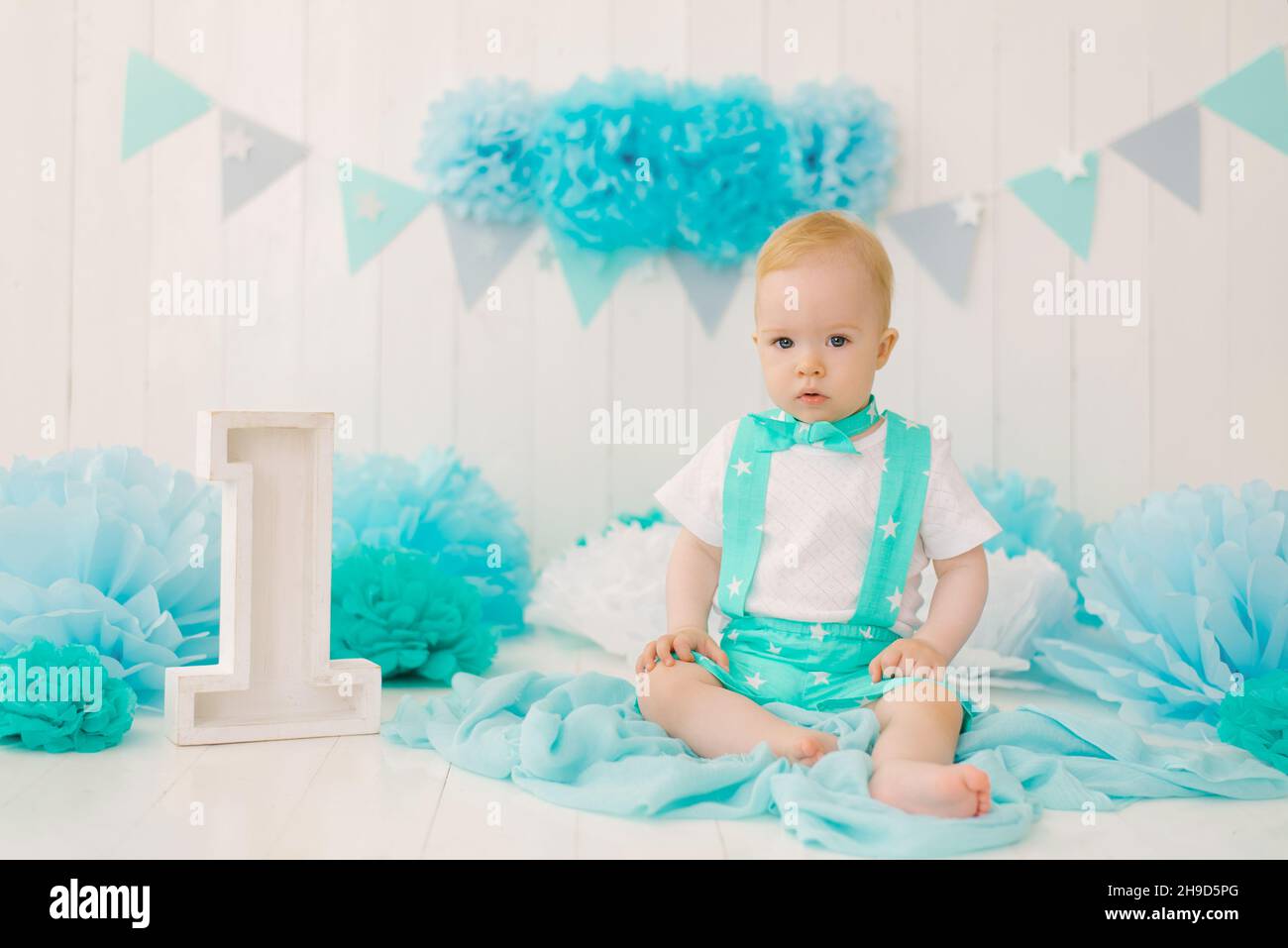 Un beau garçon d'un an en vêtements turquoise est assis près du numéro 1 dans la zone de photo pour son anniversaire Banque D'Images