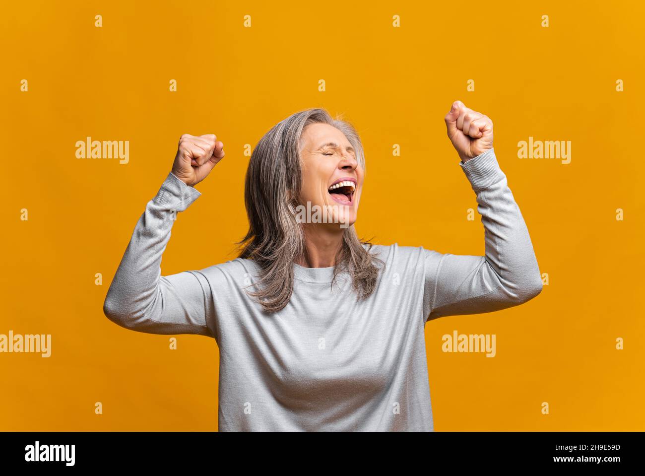 Une femme moderne mûre excitée et surjoyeuse, qui célèbre la réalisation de ses objectifs, élève des poings isolés sur une femme jaune à cheveux gris, se réjouit, criant oui dans l'euphorie, la victoire et la victoire Banque D'Images