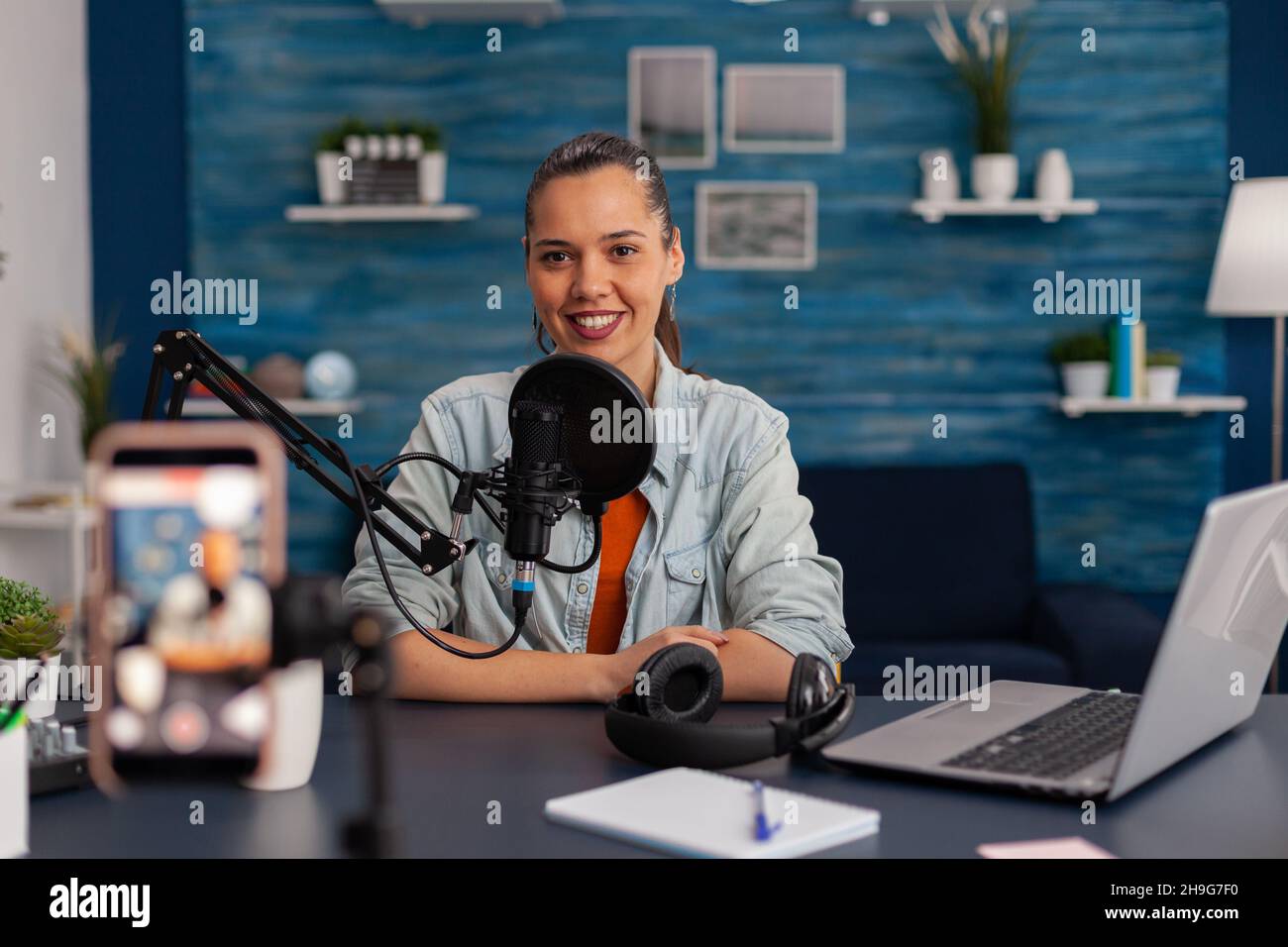 Le créateur de contenu parle devant le smartphone d'enregistrement.Un influenceur souriant diffuse des fichiers vlog sur les réseaux sociaux dans un studio à domicile.Femme diffusant un podcast avec microphone et téléphone portable. Banque D'Images