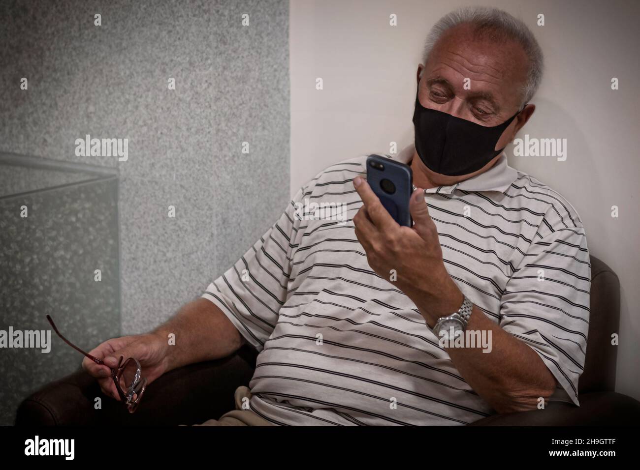 Un 70+ âgé avec des lunettes est assis dans une chaise et regarde le téléphone.Un vieil homme attend son tour à l'hôpital.Retraite Banque D'Images