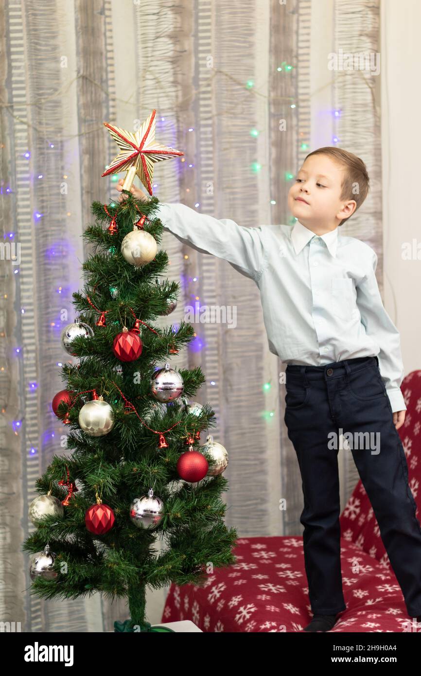 Un garçon de sept ans dans une chemise décorera un arbre de Noël avec des jouets à la veille du nouvel an à la maison sur fond de guirlandes.Mise au point sélective.Port Banque D'Images