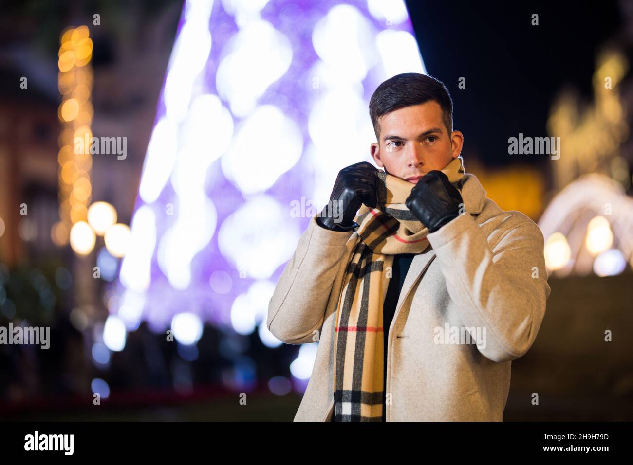 Jeune homme ajustant son foulard en hiver dehors.Arbre de Noël en arrière-plan. Banque D'Images