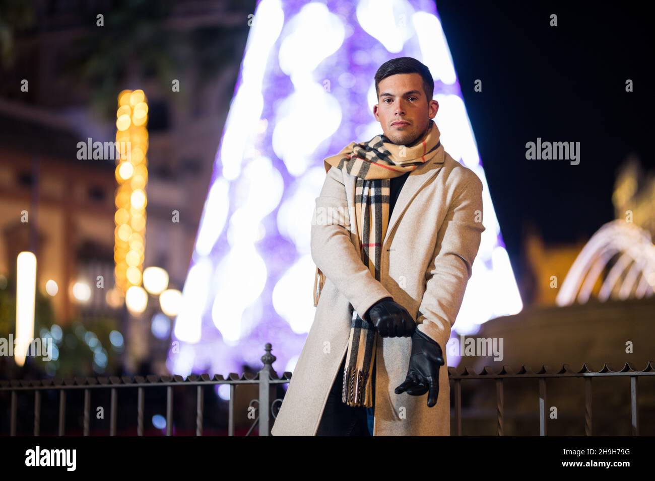 Jeune homme ajustant ses gants la nuit avec un foulard en hiver.Arbre de Noël en arrière-plan. Banque D'Images