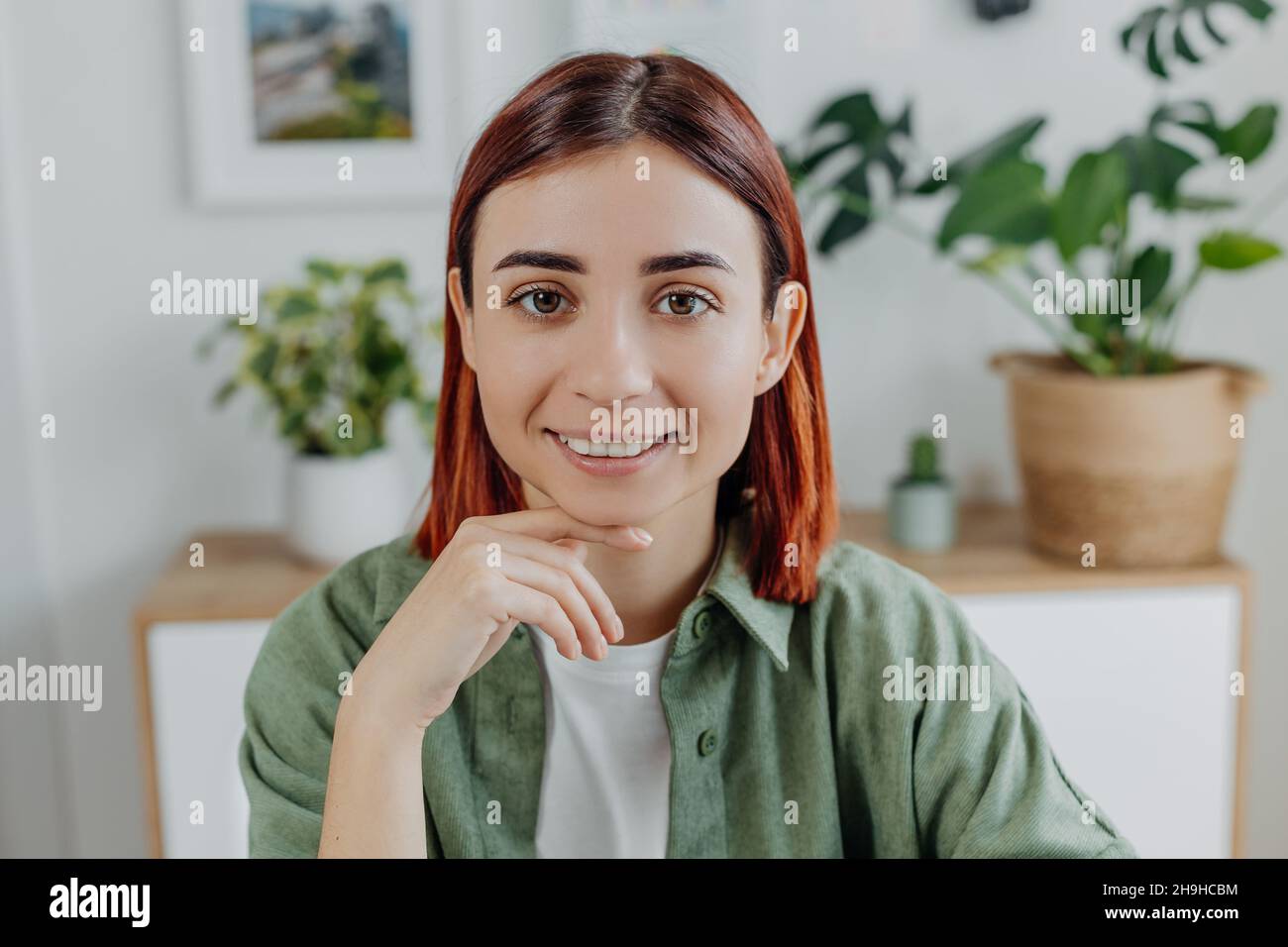 Portrait en gros plan d'une jeune femme souriante à tête rouge à la maison.Fille adulte en t-shirt vert sur le fond des plantes de maison.Concept de style de vie, positif Banque D'Images