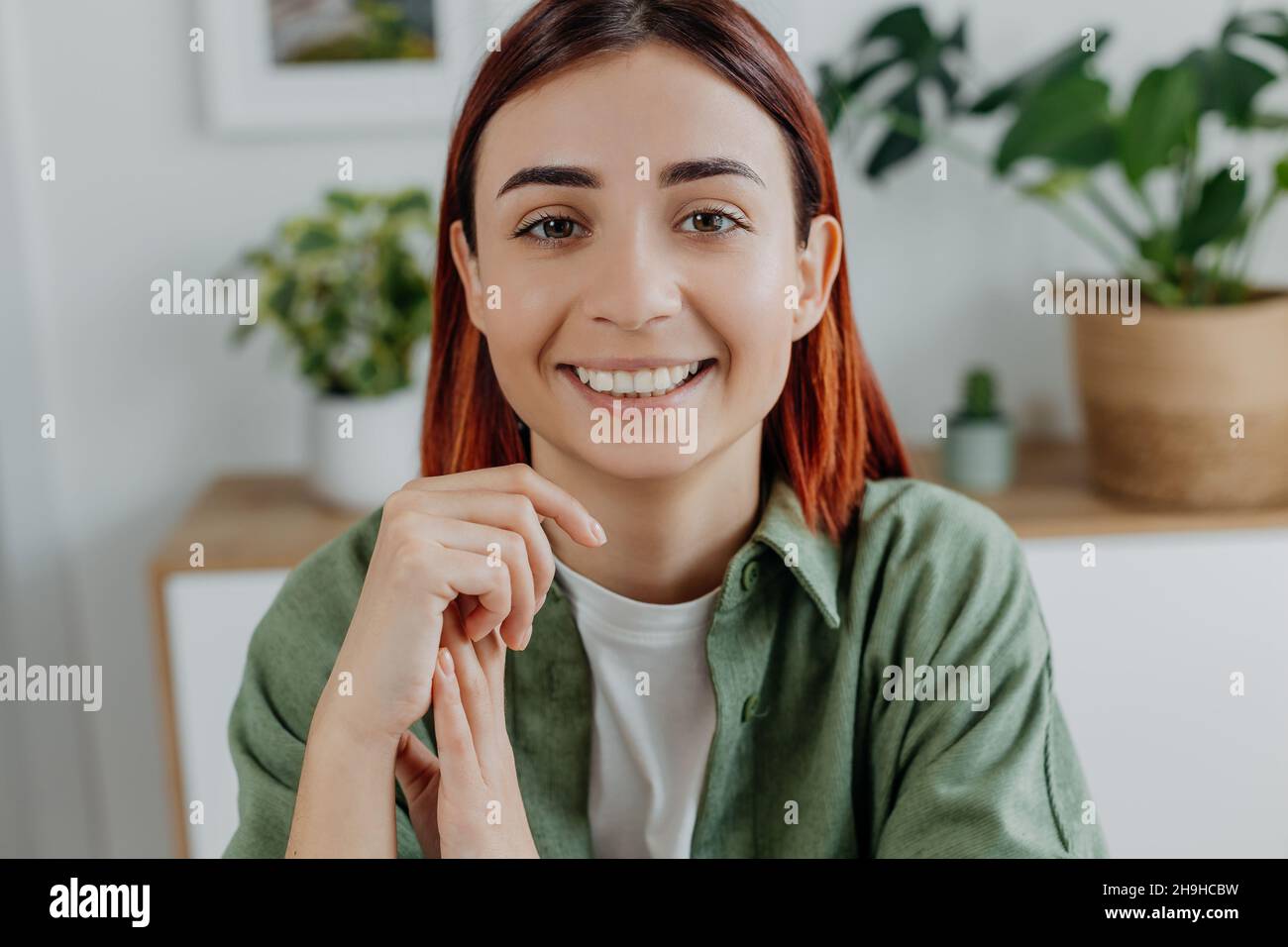 Portrait en gros plan d'une jeune femme souriante à tête rouge à la maison.Fille adulte en t-shirt vert sur le fond des plantes de maison.Concept de style de vie, positif Banque D'Images