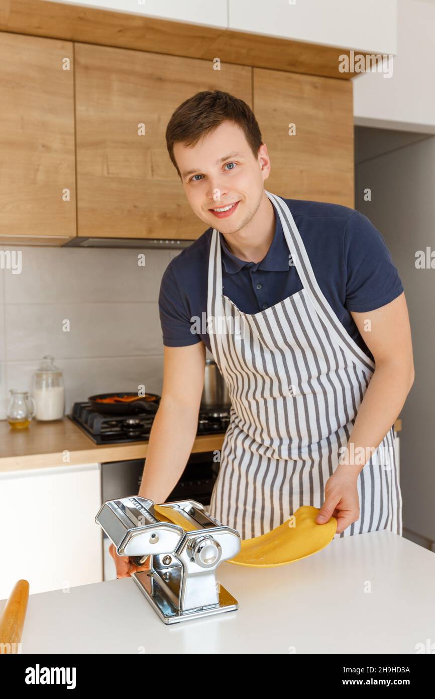 Jeune homme beau dans un tablier roulant de pâte dans la cuisine  moderne.Chef cuisant des pâtes avec machine à couper les pâtes.Concept de  la nourriture maison, cuisine masculine et s Photo Stock -