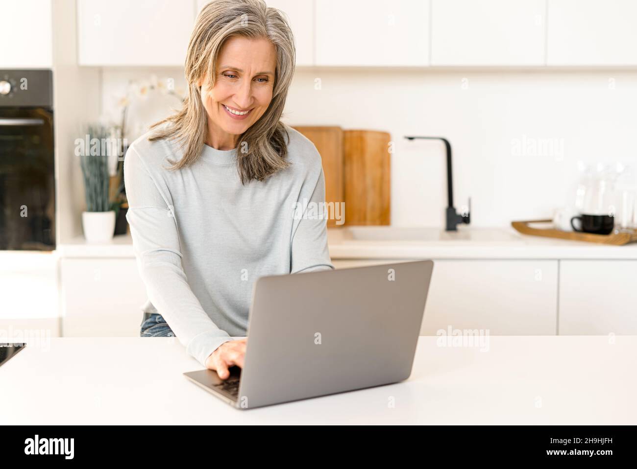 Positive femme âgée s'asseoir se détendre dans la cuisine, travailler sur ordinateur portable, sourire, moderne mature femme des années 60 profiter d'un week-end de loisirs à la maison, la navigation, le Web surf sur ordinateur, shopping, la saisie de message Banque D'Images