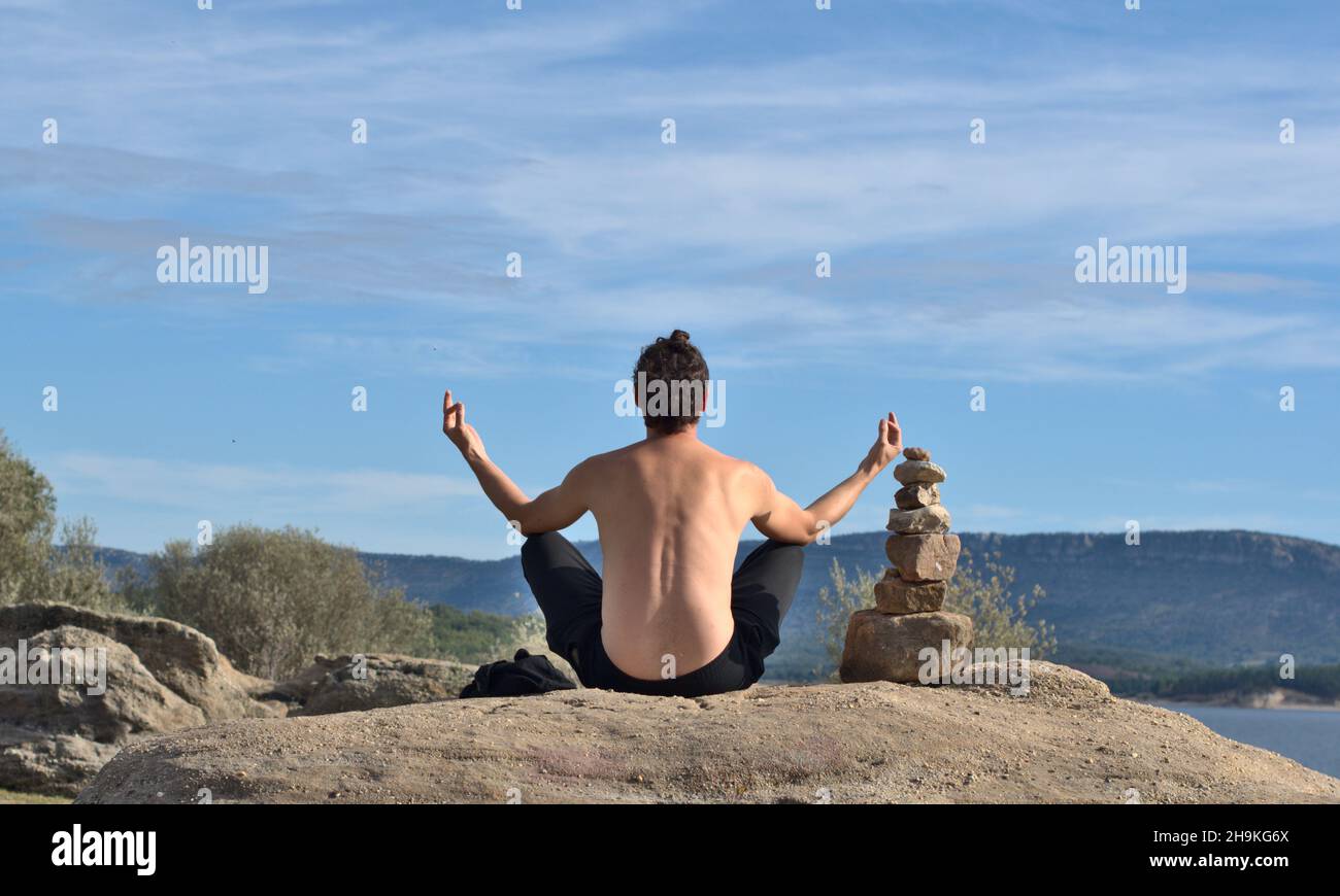 Homme de derrière avec torse nu faisant du yoga, les doigts en position mudra.Pile de pierres à côté de l'homme, symbole de l'équilibre.Lac et montagne avec PIN Banque D'Images