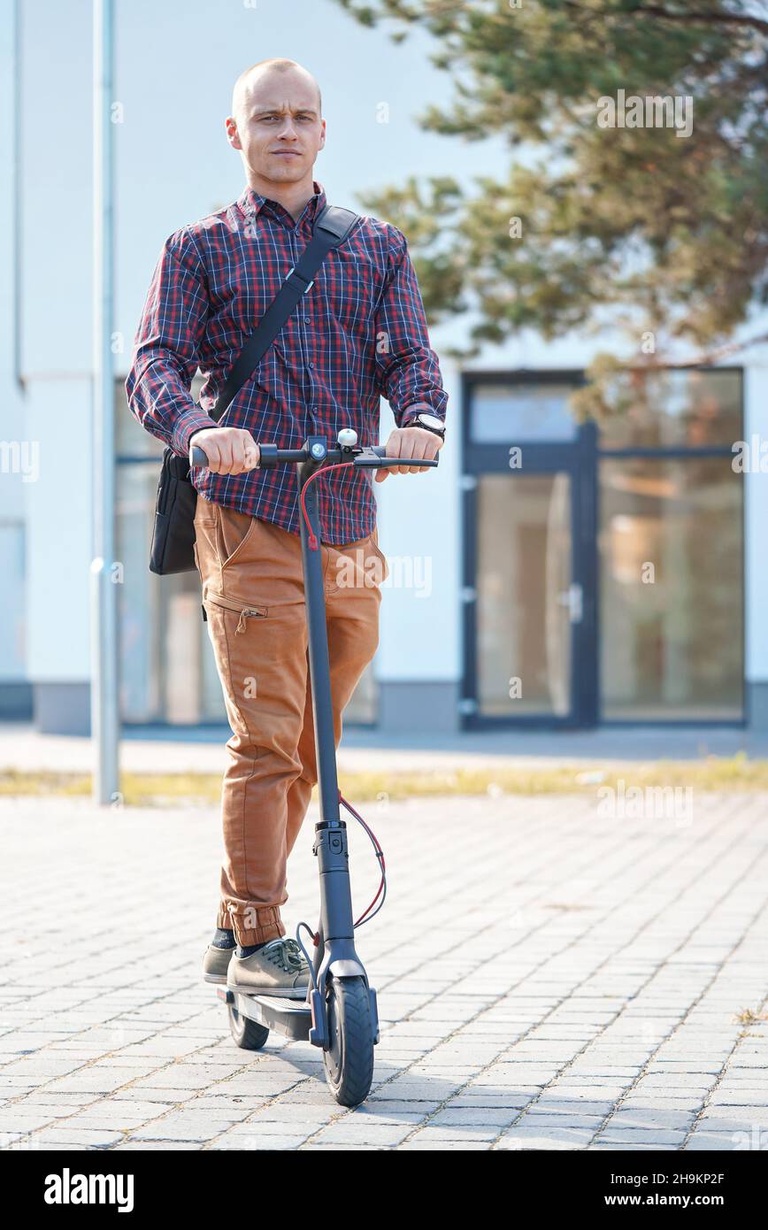 Jeune homme dans un pantalon décontracté et une chemise à cheval sur son scooter électrique, vue de face, bâtiments à l'arrière-plan Banque D'Images