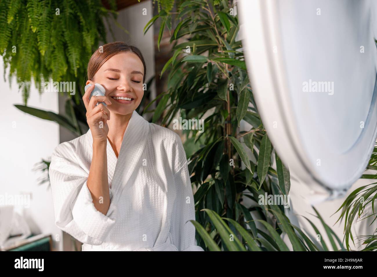 Bonne jeune femme en peignoir souriant et appréciant l'utilisation de la brosse en silicone pour la peau exfoliante, debout dans la salle de bains décorée de vert Banque D'Images