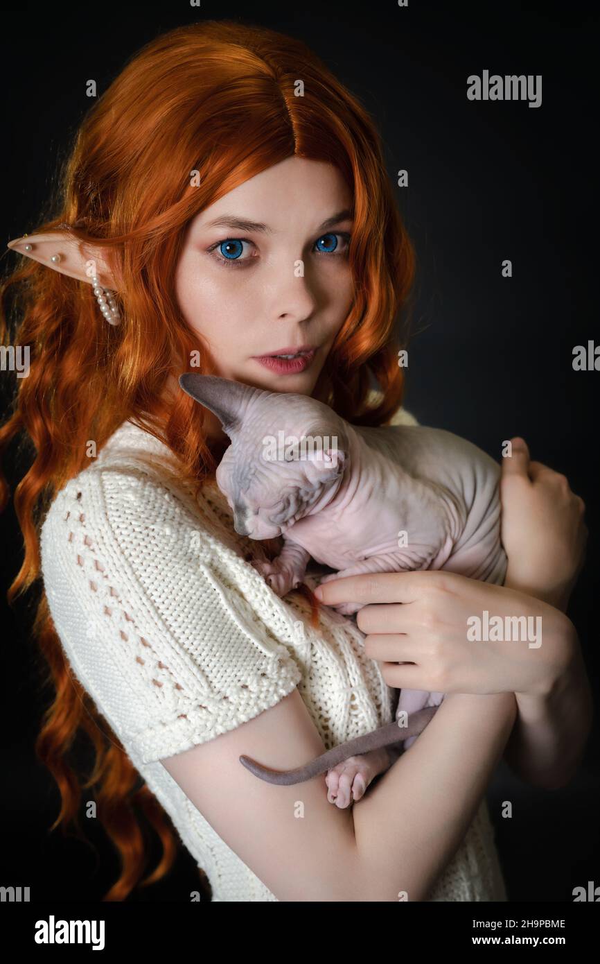 Portrait d'une jeune femme cosplayer elf, embrassant le chaton de Sphinx.Elf en robe tricotée blanche, avec de luxueux cheveux longs rouges et des oreilles percées Banque D'Images