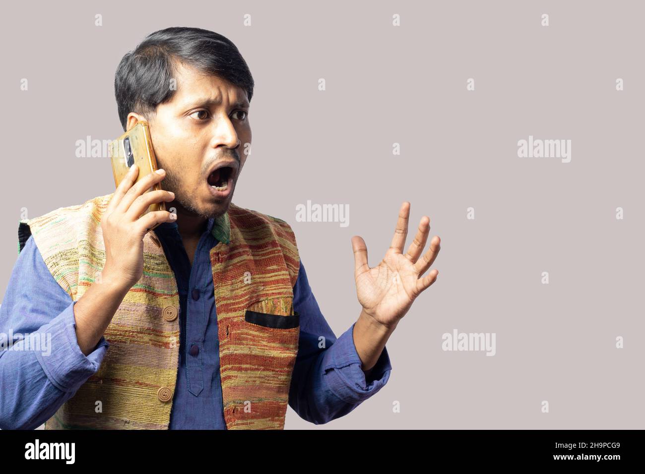 Un jeune Indien en robe bleue parlant au téléphone avec expression agitée sur fond gris Banque D'Images