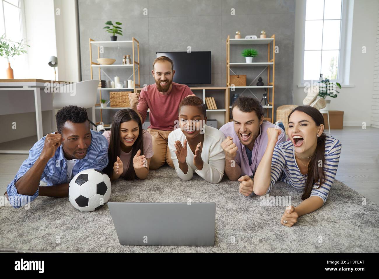 Des amis se trouvent sur le sol dans le salon, devant un ordinateur portable qui diffuse un match de football. Banque D'Images