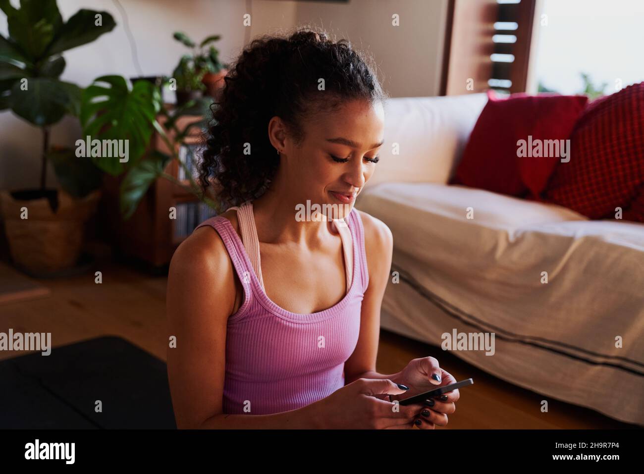 Happy Multi-culture jeune femme adulte dactylographiant sur son téléphone tout en étant sur le tapis de yoga dans le salon moderne à la maison Banque D'Images