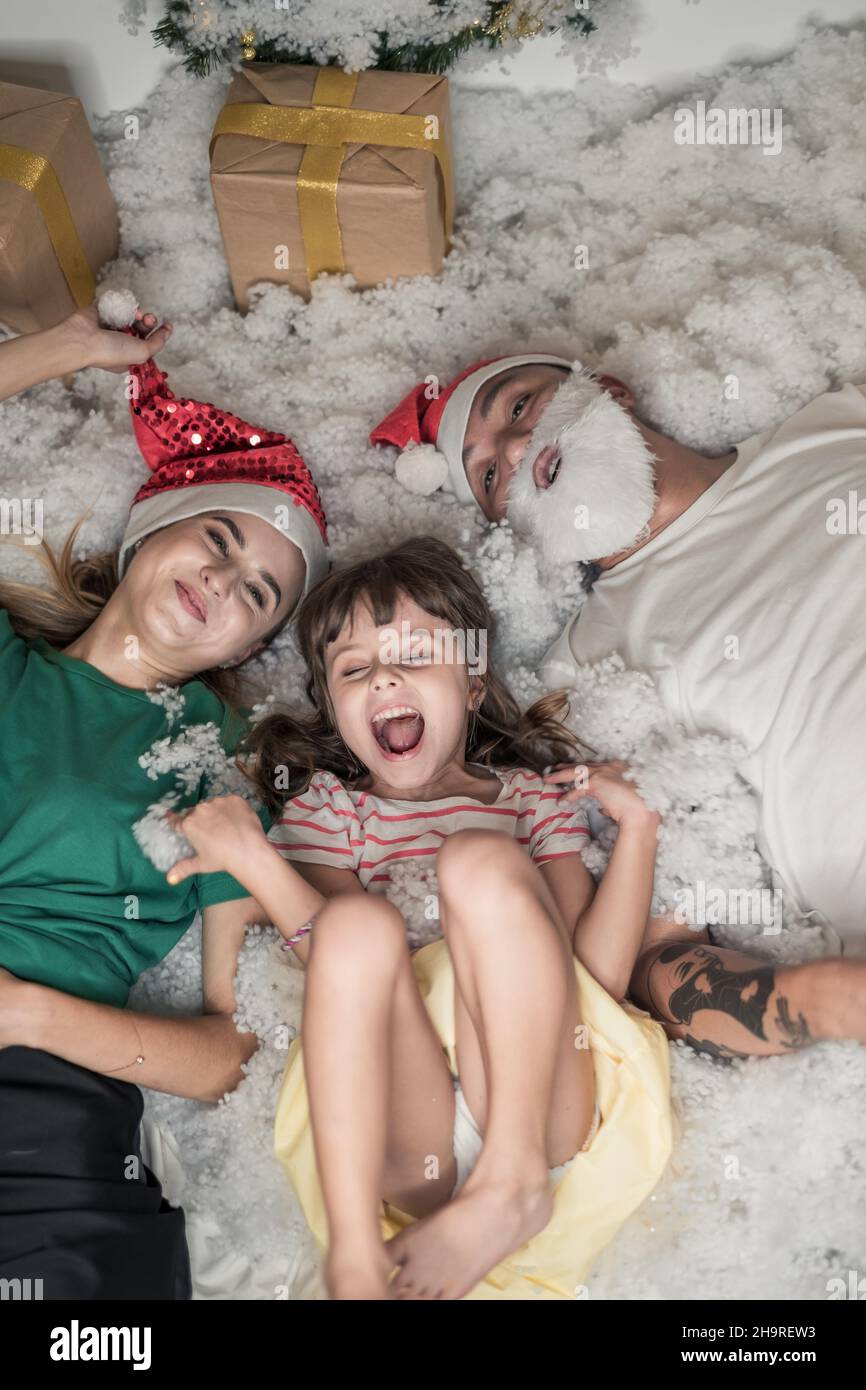 Photo de Noël de la famille heureuse.Nouvel an Portrait de papa, maman et fille portant des chapeaux rouges de Noël, souriant et allongé dans la neige artificielle blanche. Banque D'Images