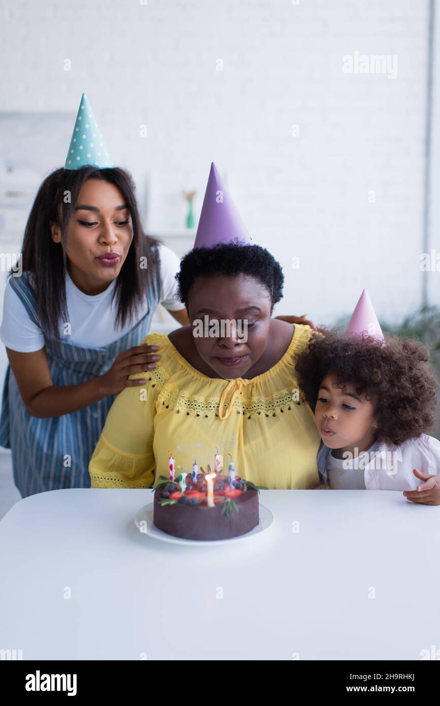 afro-américaines femmes et petite fille dans des casquettes de fête soufflant des bougies sur le gâteau d'anniversaire Banque D'Images