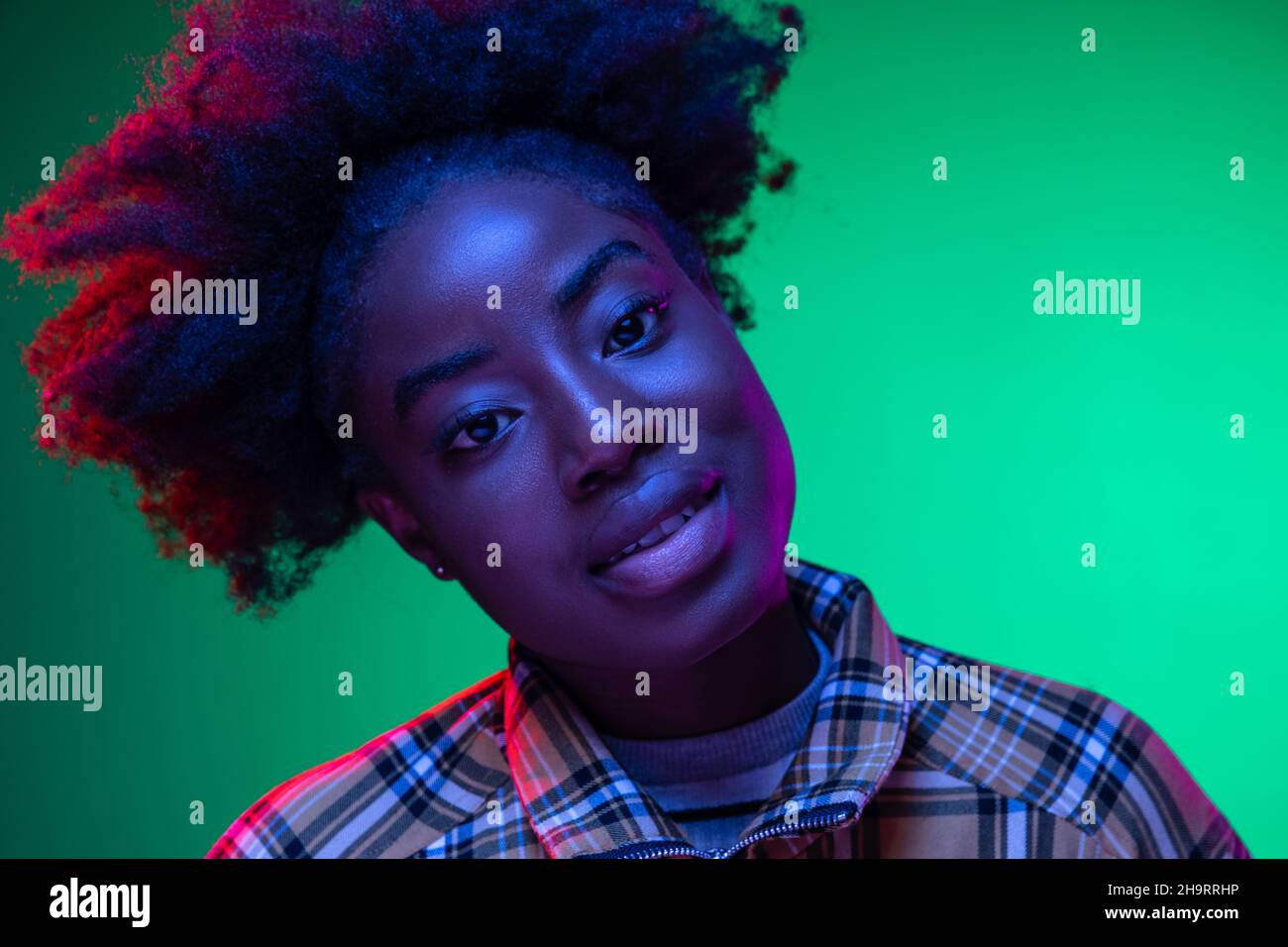 Gros plan sur le portrait d'une jeune fille africaine sur fond de studio vert foncé, dans une lumière de néon mauve.Hairdo afro.Concept des émotions humaines Banque D'Images