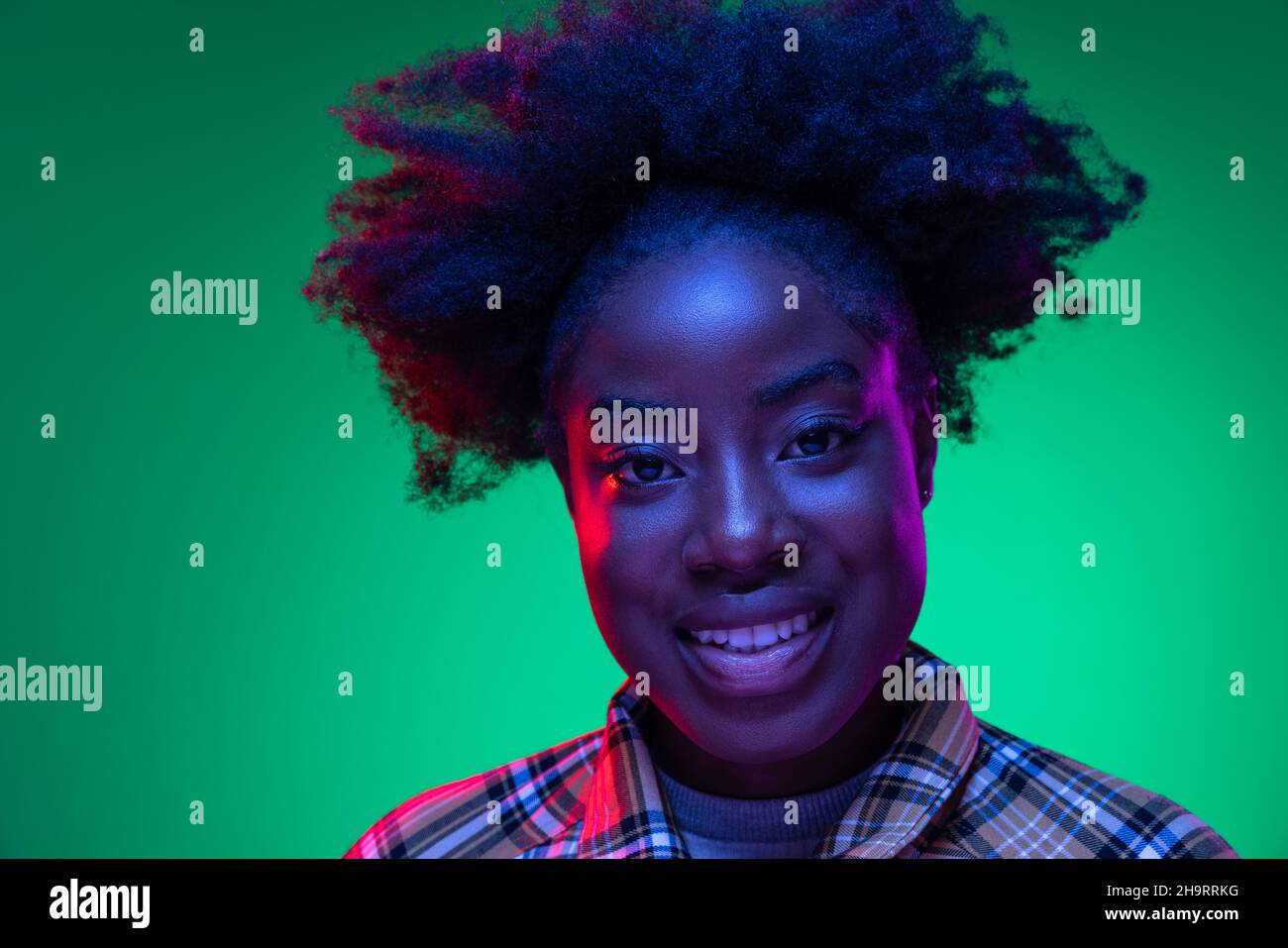 Gros plan sur le portrait d'une jeune fille africaine sur fond de studio vert foncé, dans une lumière de néon mauve.Hairdo afro.Concept des émotions humaines Banque D'Images