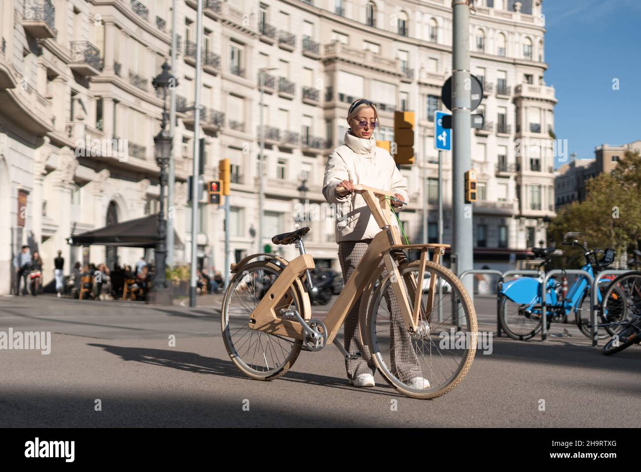 Femme élégante avec vélo écologique debout sur la route asphaltée à l'extérieur de l'immeuble d'appartements le jour ensoleillé dans le centre-ville. Banque D'Images