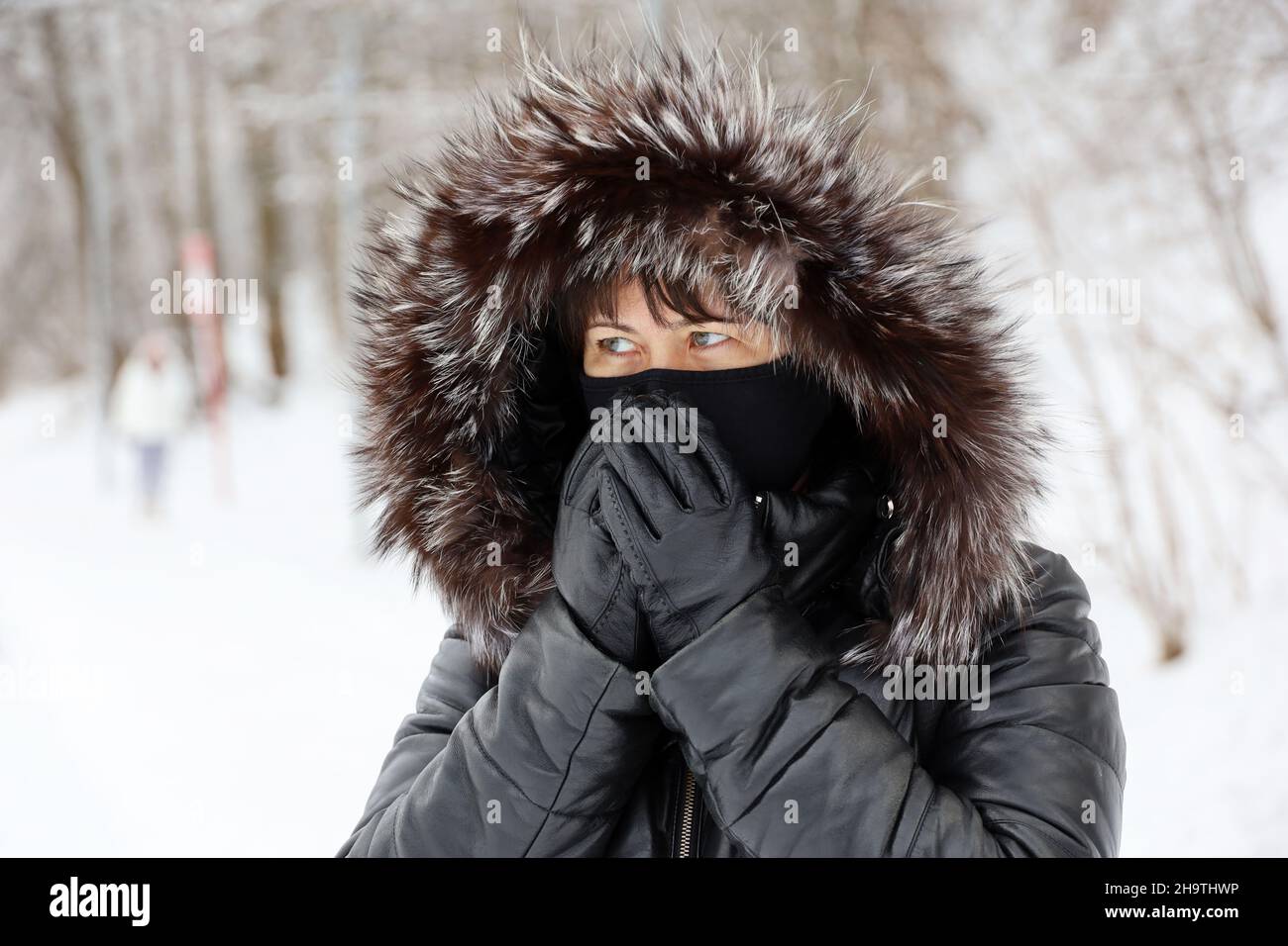 Femme en masque noir et capuche en fourrure debout dans une rue pendant la neige et couvrant son visage par les mains dans des gants en cuir.Temps froid en ville d'hiver Banque D'Images
