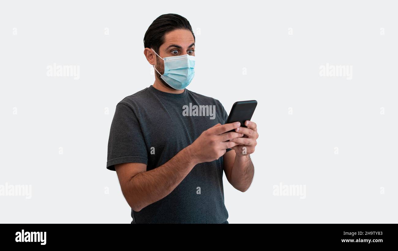 Portrait d'un homme caucasien qui soulève des sourcils en raison de la surprise avec un smartphone et un masque médical pendant l'épidémie de virus corona.Réaction surprise perso Banque D'Images