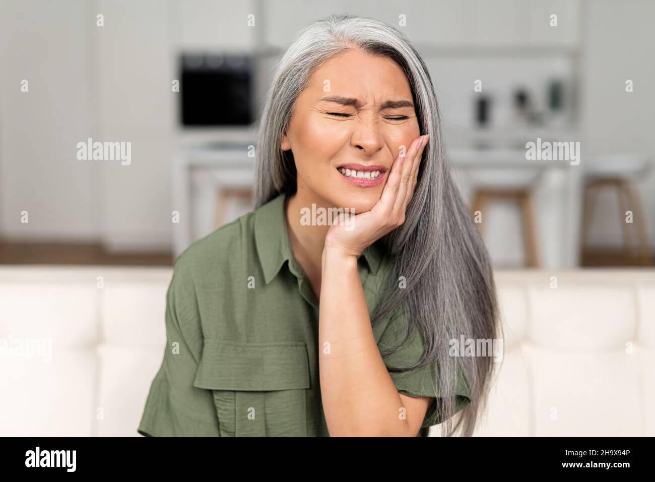 Trouble mature 50s femme avec cheveux gris se sent mal de dents fort, tenant la joue et souffrant de douleur dentaire forte assis sur le canapé, moyenne-âge femelle subissant des douleurs dentaires, sensibilité à l'émail Banque D'Images