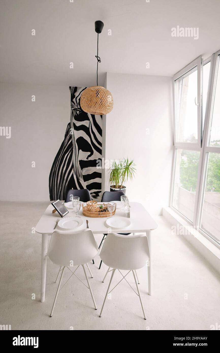 Décoration intérieure d'une salle à manger lumineuse dans un style minimaliste avec une table blanche en bois carré, chaises noires et blanches, avec des décorations en osier sur le lustre et sur la table, avec une grande fenêtre et une énorme image de zèbre sur le mur.Photo de haute qualité Banque D'Images