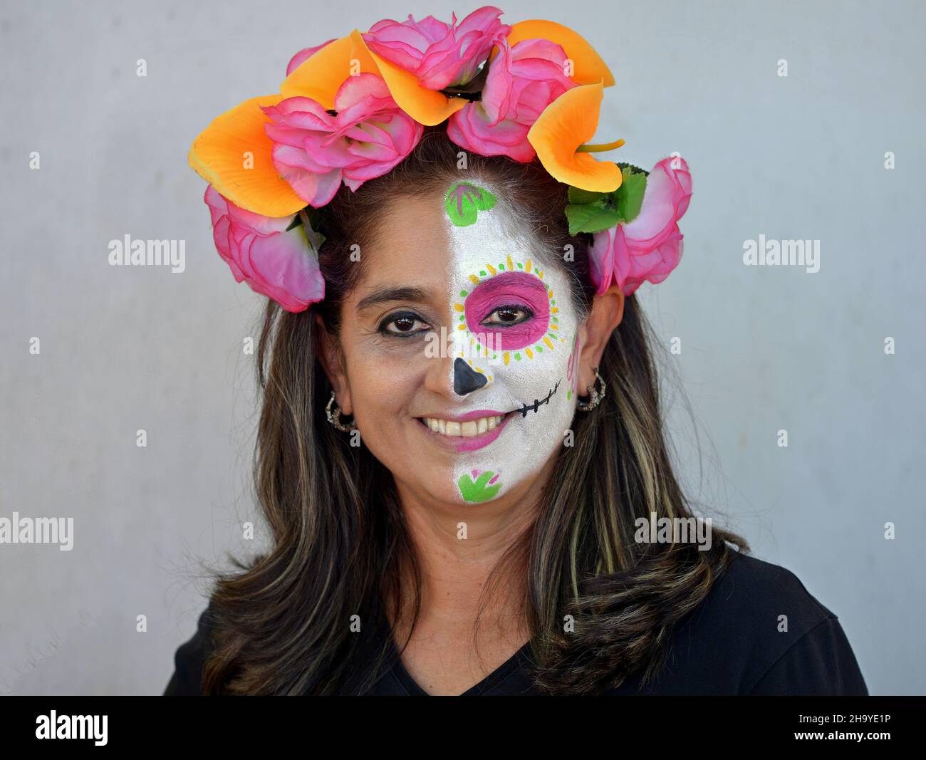 Une mexicaine aux fleurs colorées dans les cheveux peint la moitié gauche de son visage avec Catrina visage maquillage le jour des morts et sourit au spectateur. Banque D'Images