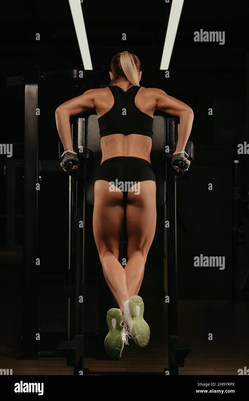 Une photo de derrière une femme en forme avec des cheveux blonds qui fait des push-up sur les barres inégales dans une salle de gym.Une jeune fille forme ses bras et sa poitrine. Banque D'Images