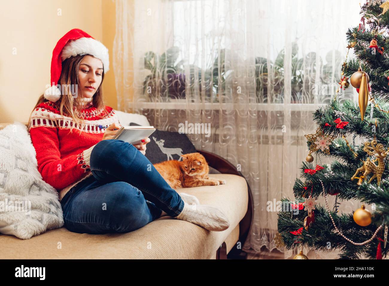 Jeune femme écrivant la liste des objectifs pour 2022 nouvel an dans le carnet à la maison assis par arbre de Noël portant chapeau de père Noël et chandail chaud. Banque D'Images