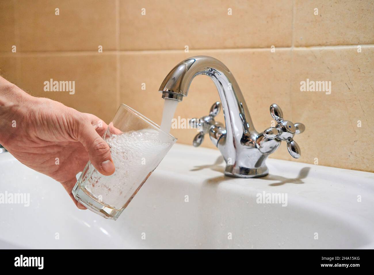 Homme remplissant un verre d'eau à partir d'un robinet à colonne en acier inoxydable.La main d'un homme verse de l'eau dans le verre du robinet chromé de la salle de bains pour boire de l'eau courante Banque D'Images
