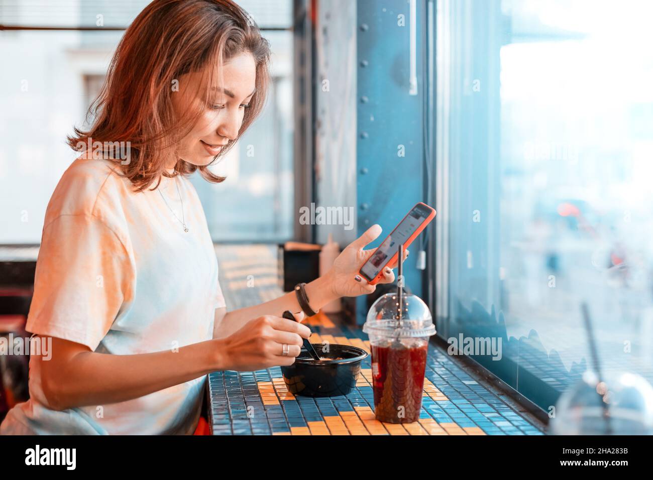 Une femme asiatique mange dans une cantine de restauration rapide et envoie des SMS à un ami via un messager sur son smartphone Banque D'Images