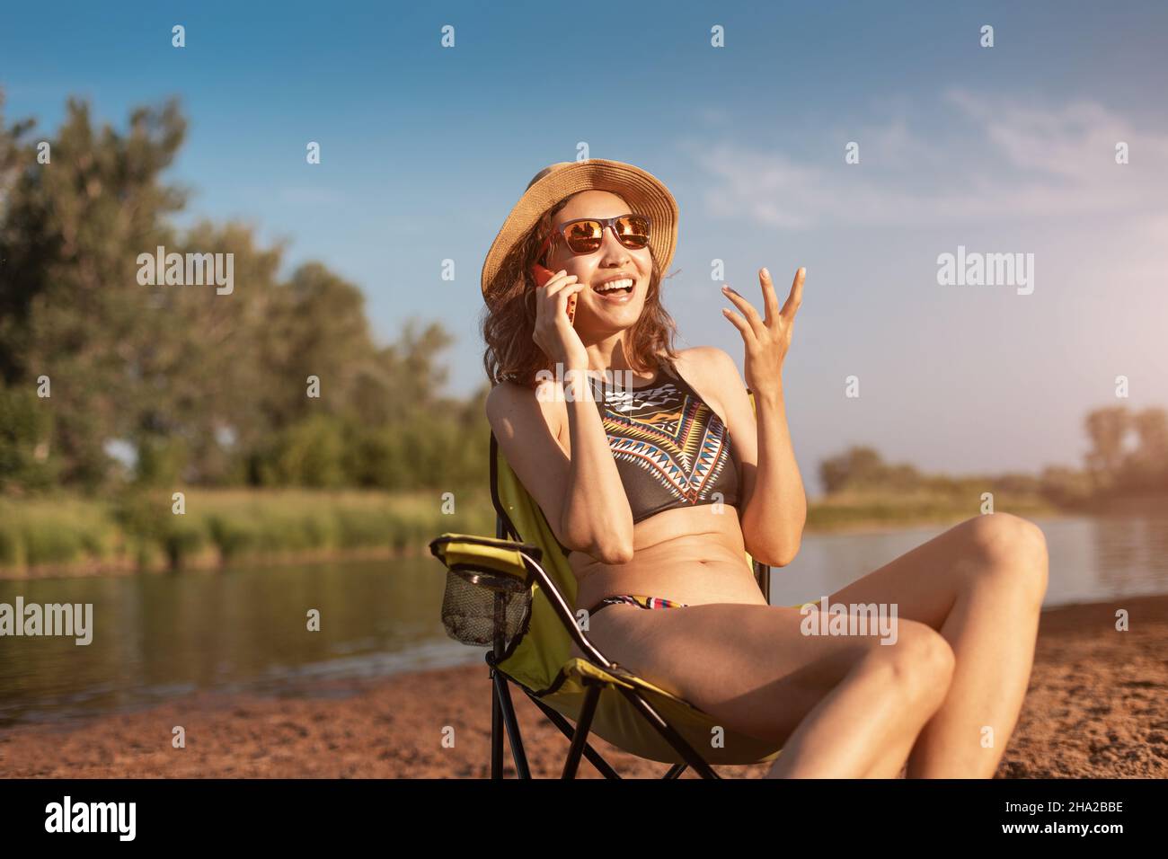 Une femme heureuse communique avec un ami sur un smartphone tout en étant assise dans une chaise de camping sur la rive de la rivière.Vacances d'été par l'eau et mobile Inter Banque D'Images